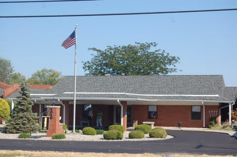 Single-story red brick building with a covered entrance, an American flag on a pole, and landscaped shrubs in front.