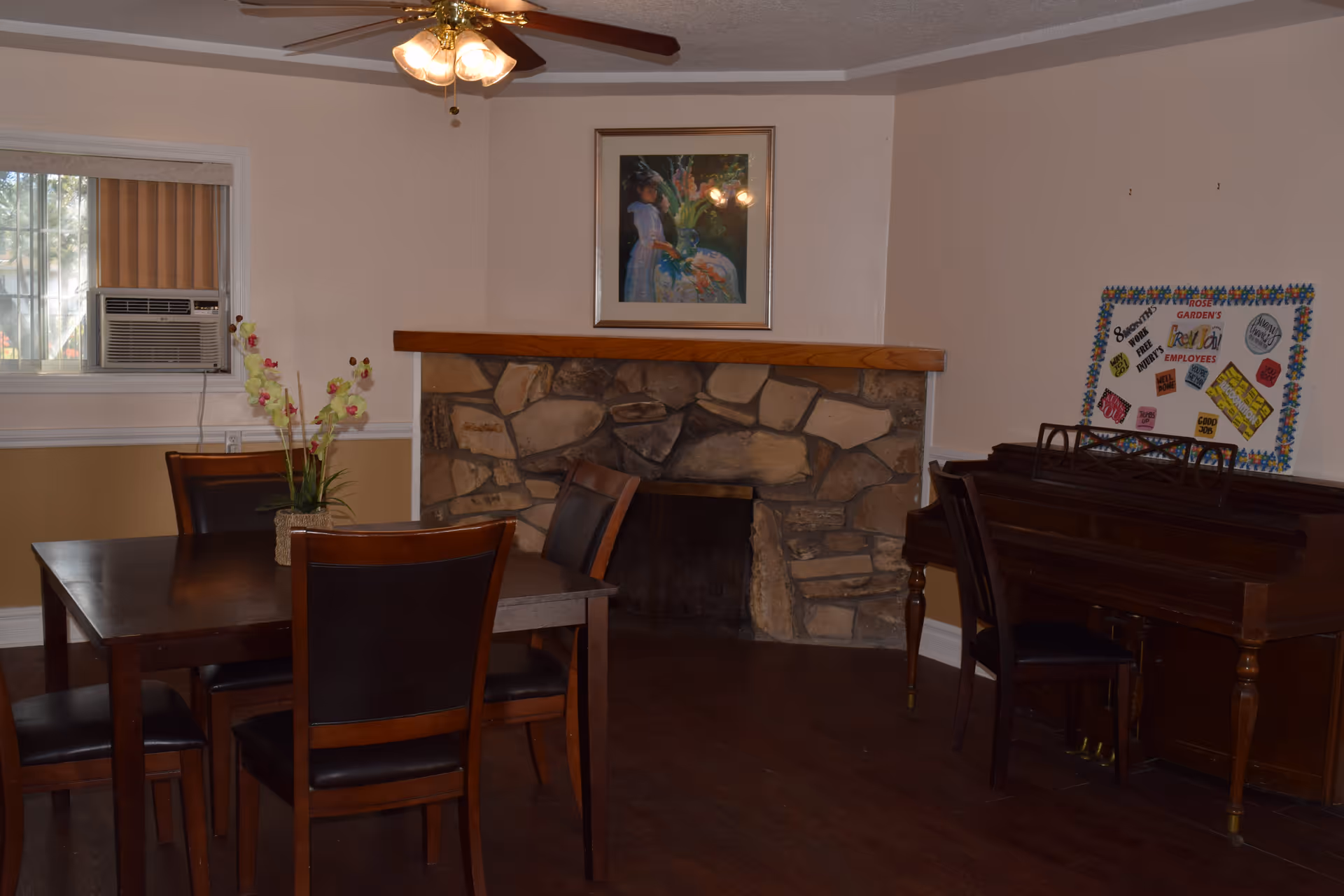 A cozy room with a wooden dining table and four chairs, a stone fireplace with a wooden mantel, a framed painting above the fireplace, a piano with a chair, and a bulletin board on the wall. There is a window with an air conditioning unit and vertical blinds, and a ceiling fan with lights.
