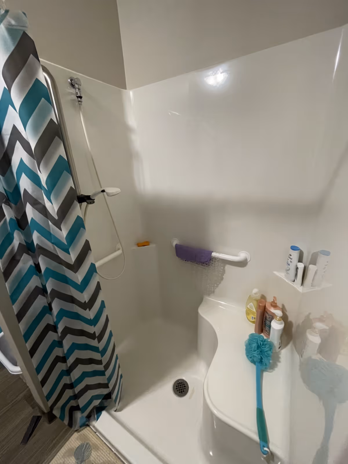 A white shower stall with a built-in seat and grab bars. There is a blue and gray chevron-patterned shower curtain partially drawn to the side. Various shower products and a blue bath scrubber are placed on the seat and built-in shelves.