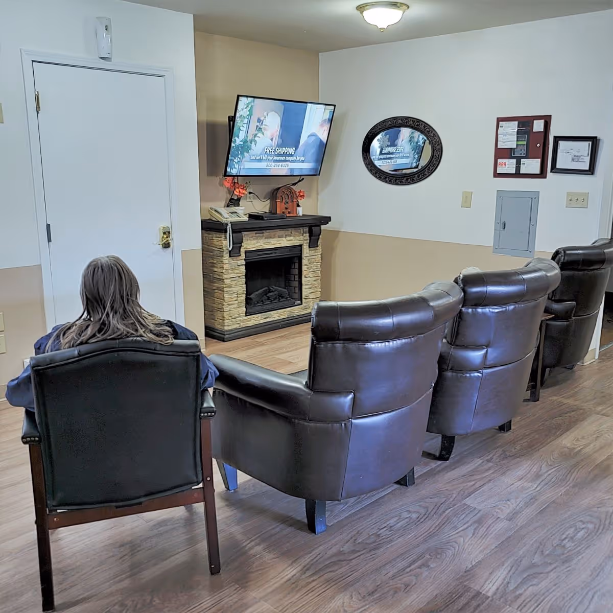 A person sitting in a black chair watching a wall-mounted TV in a room with three additional leather armchairs arranged in a row. The room has a small fireplace below the TV, a round decorative mirror on the wall, and a bulletin board with papers. The floor is wooden, and the walls are painted white and beige.