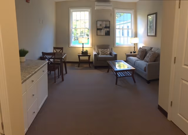 A cozy living room area in Beaver Lake Lodge Assisted Living featuring a gray sofa with patterned cushions, a matching armchair, a glass-top coffee table, two side tables with lamps, and two large windows letting in natural light. There is also a small dining table with chairs and a white cabinet with a granite countertop on the left side.