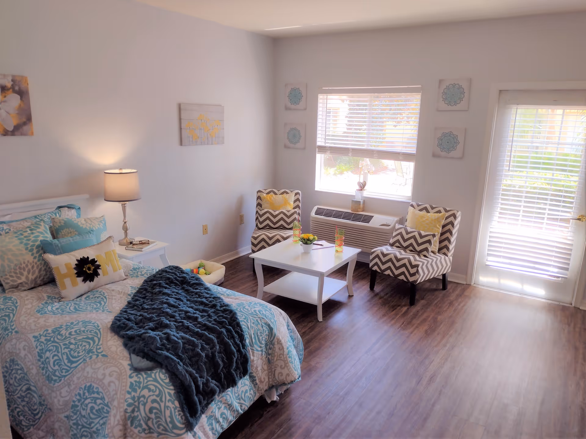 A bright and cozy bedroom with a bed covered in blue and white patterned bedding, a dark blue throw blanket, and a decorative pillow that says 'HOME'. Next to the bed is a white nightstand with a lamp and a book. Across from the bed are two chevron-patterned chairs with yellow pillows, a white coffee table with two colorful glasses and a small plant, and a window with blinds letting in natural light. There is also a door with blinds leading outside, and the floor is a dark wood laminate.