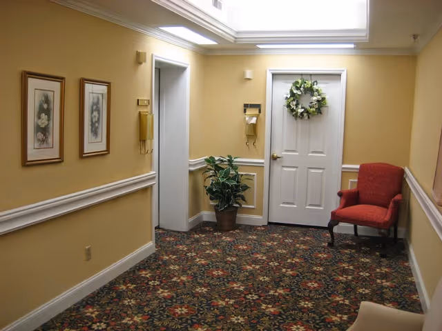 A hallway with yellow walls and floral carpet. There are two framed floral paintings on the left wall, a potted plant near a white door decorated with a green wreath, and a red upholstered chair on the right side. The ceiling has recessed lighting.