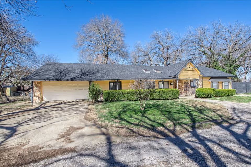 Single-story yellow house with an attached two-car garage, trimmed hedges and a front lawn under a clear blue sky.