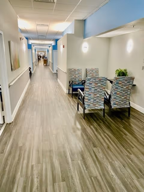 A long, well-lit hallway with wood-style flooring and light-colored walls. On the right side, there is a small seating area with four patterned chairs around a table and a small green plant. The hallway extends into the distance with doors and artwork visible along the walls.