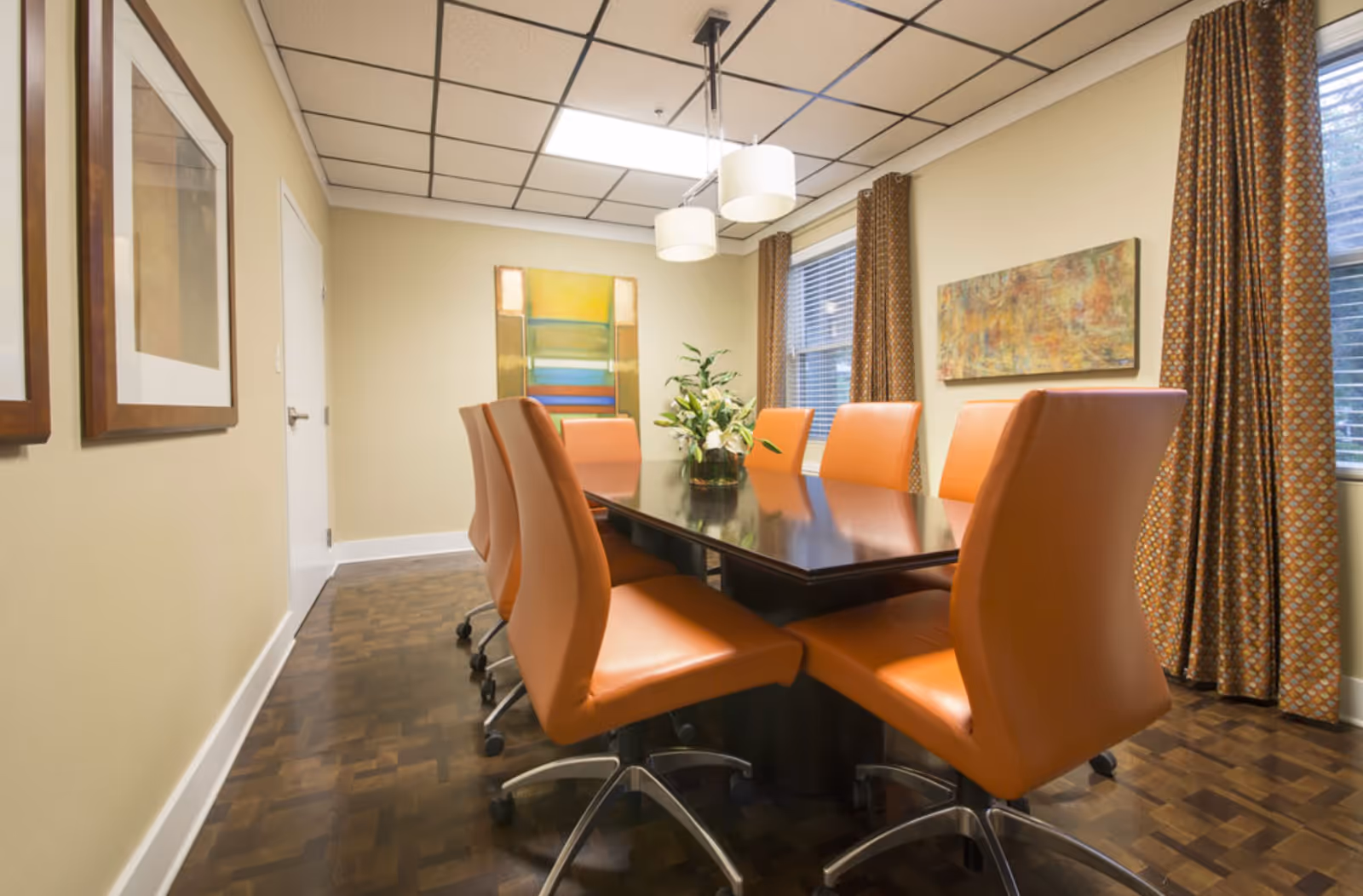 A modern dining/meeting room with a dark wood table surrounded by orange swivel chairs, artwork on the walls, and pendant lights overhead.