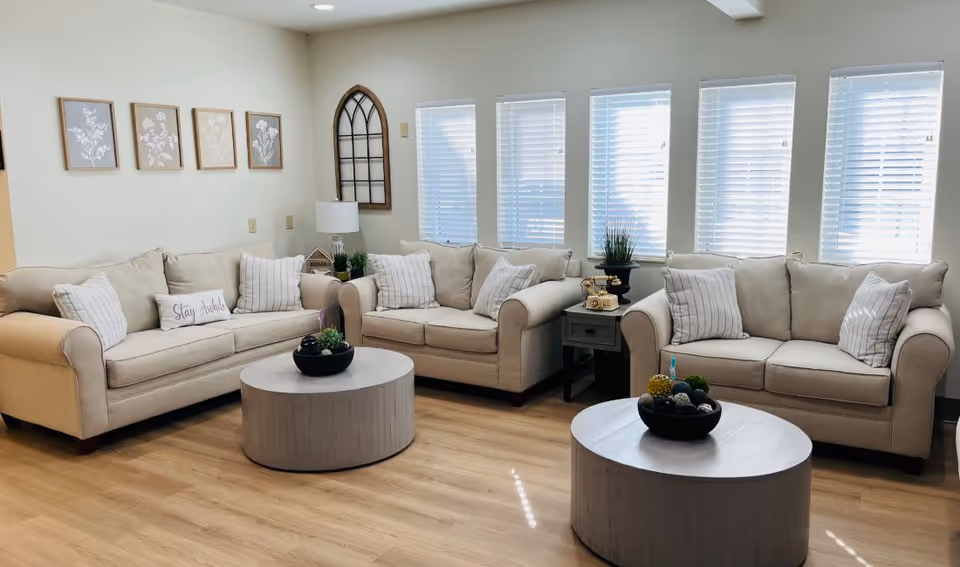 A bright living room with three beige sofas adorned with striped and solid pillows. Two round wooden coffee tables with decorative bowls are placed in front of the sofas. The room has light wood flooring, five tall windows with white blinds, and wall decorations including framed botanical prints and a windowpane-style mirror. A small side table with a vintage rotary phone and a lamp is positioned between two sofas.