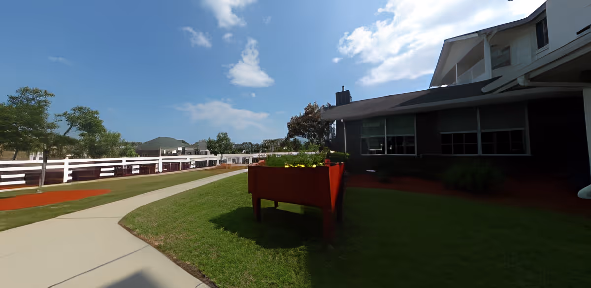 Outdoor view of a senior living facility with a paved walkway, green grass, trees, and a raised red planter box with flowers. The building is partially visible on the right side under a blue sky with some clouds.