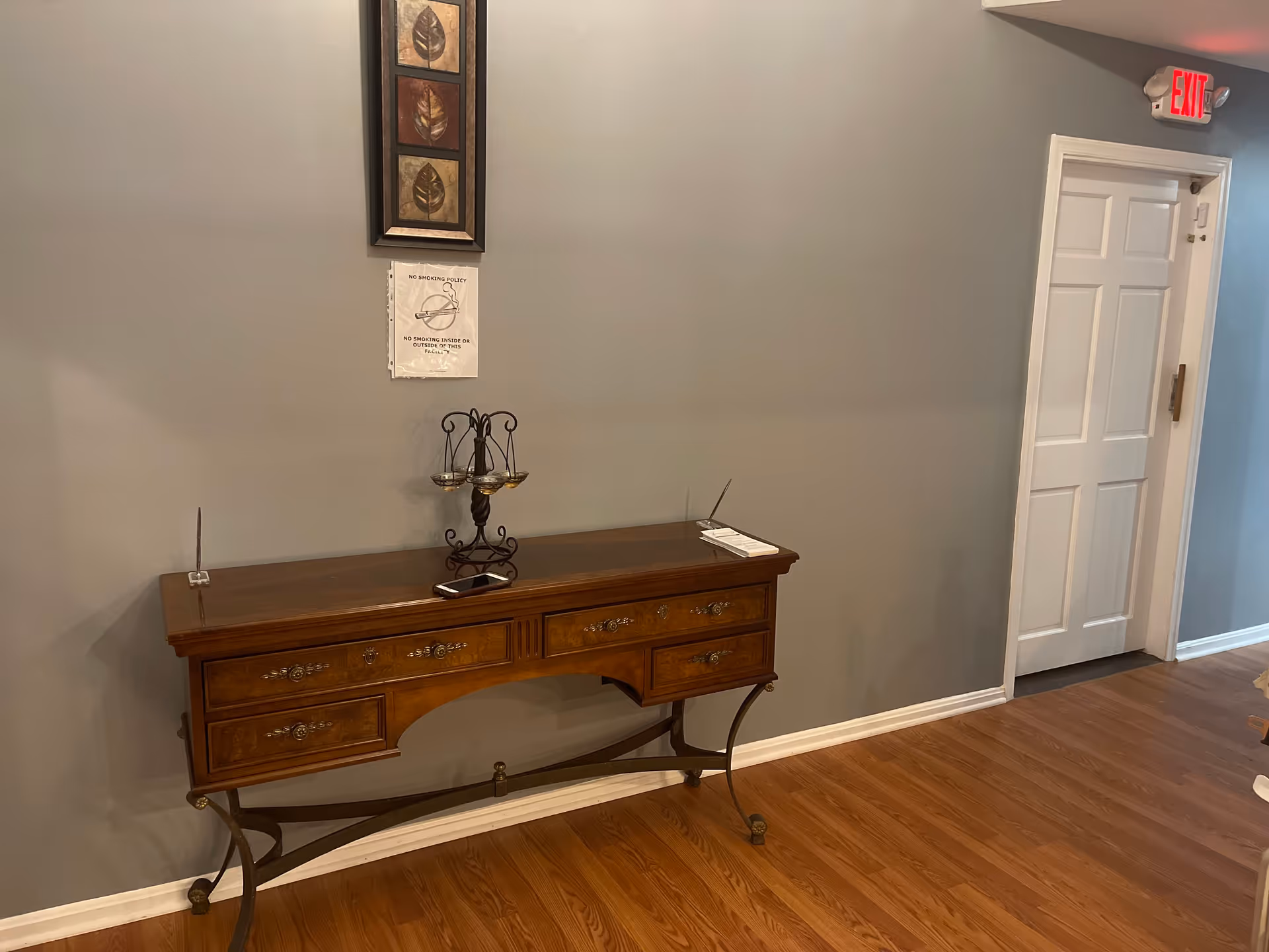A hallway with wooden flooring and gray walls featuring a wooden console table with decorative metal legs. On the table are a small decorative item and a smartphone. Above the table is a framed artwork with leaf designs and a no smoking policy sign. A white door with an illuminated red exit sign above it is visible at the end of the hallway.