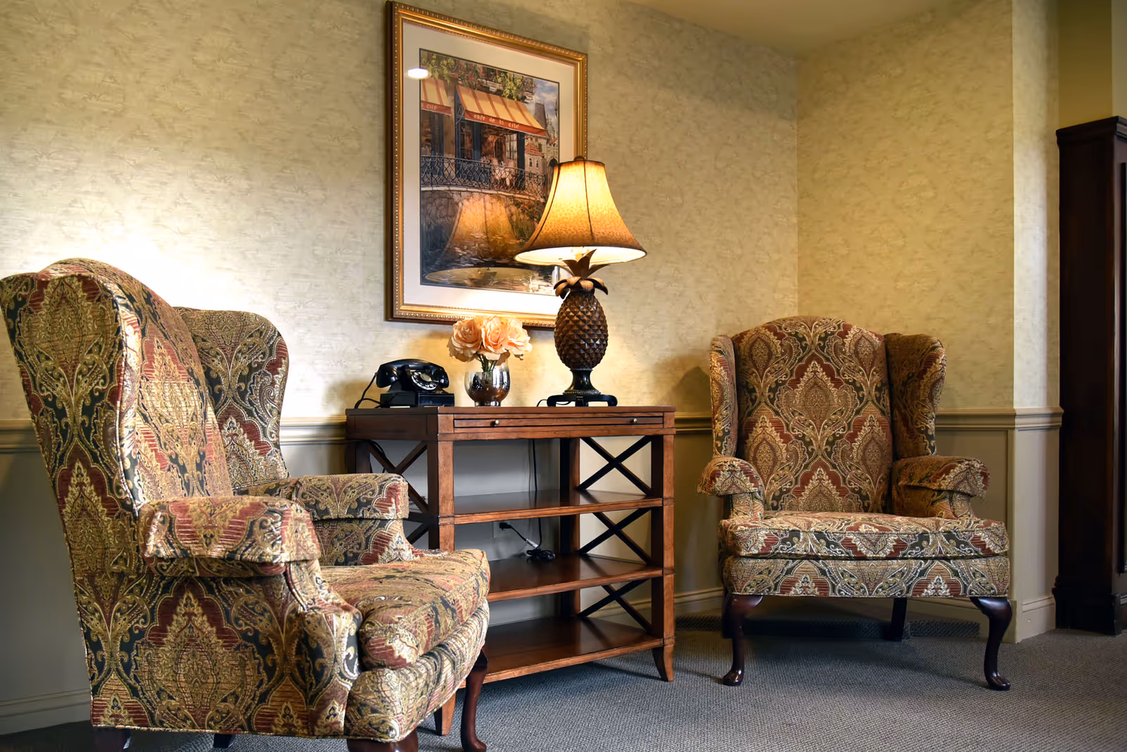 Two patterned wingback chairs flank a wooden console table topped with a lamp, telephone, and flowers beneath a framed painting in a cozy sitting area.