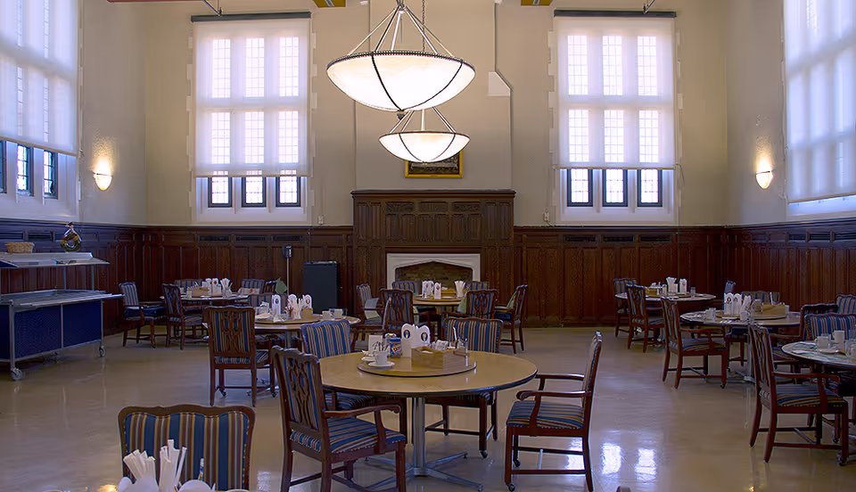 A spacious dining room with multiple round tables and wooden chairs with striped cushions. The room features large windows with white blinds, wood paneling on the walls, and two large hanging light fixtures. There is a fireplace in the center of the far wall and a serving cart on the left side.