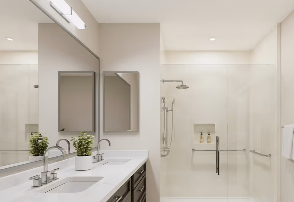 Modern bathroom with a double sink vanity featuring chrome faucets and a small potted plant. There are two rectangular mirrors above the sinks. To the right, there is a glass-enclosed walk-in shower with a rain showerhead, handheld shower, and built-in shelf holding two bottles. The walls and floor are tiled in light neutral tones, and there are grab bars inside the shower for accessibility.