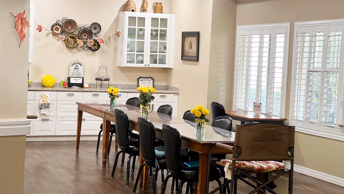 A bright dining room with a long wooden table surrounded by black chairs. The table has three vases with yellow flowers. The room features white cabinets with glass doors, decorative plates on the wall, and large windows with white shutters letting in natural light.