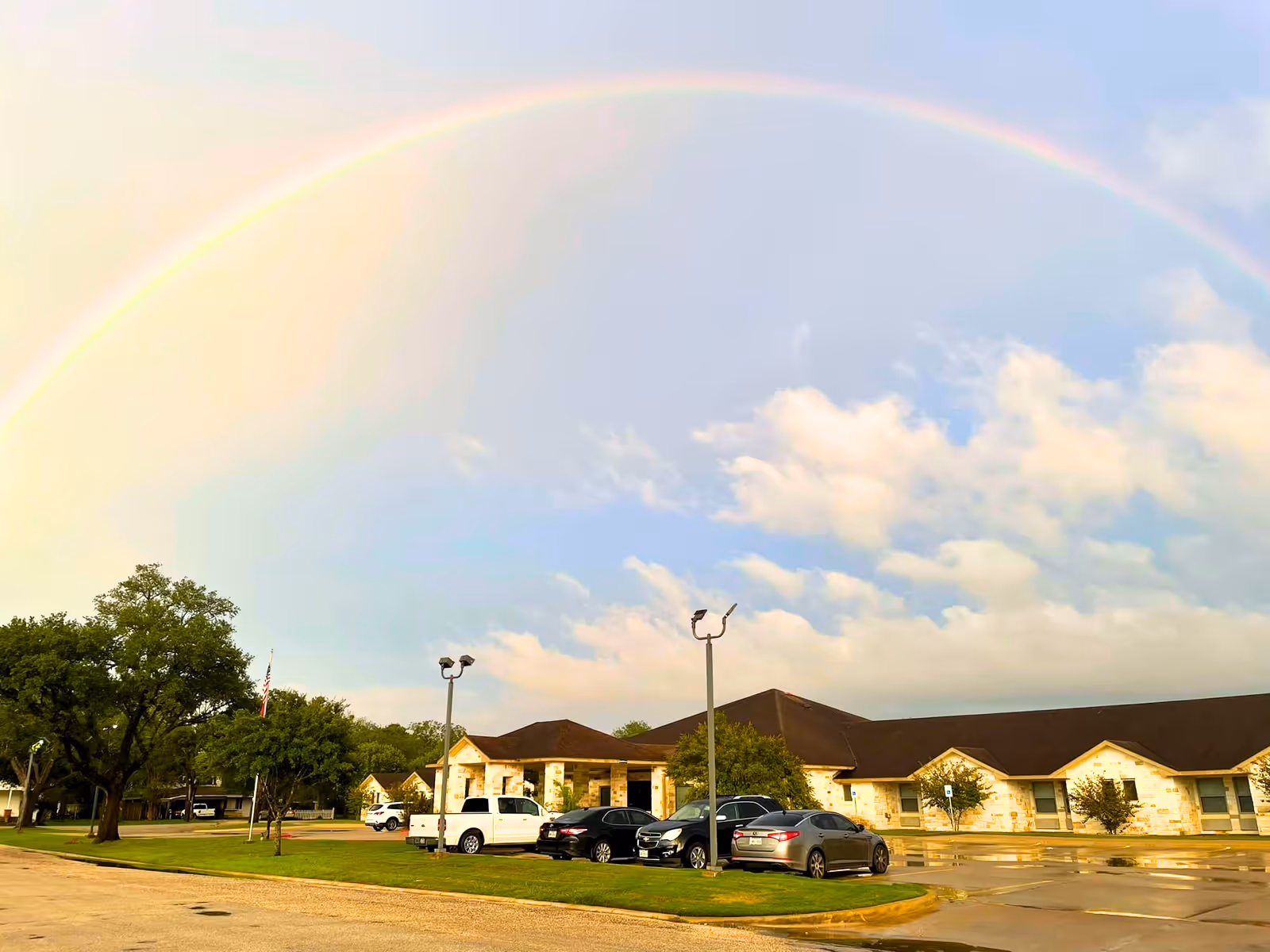 Exterior view of Ganado Nursing & Rehab Center building with a parking lot in front, several cars parked, green trees, and a rainbow arching across a partly cloudy sky.
