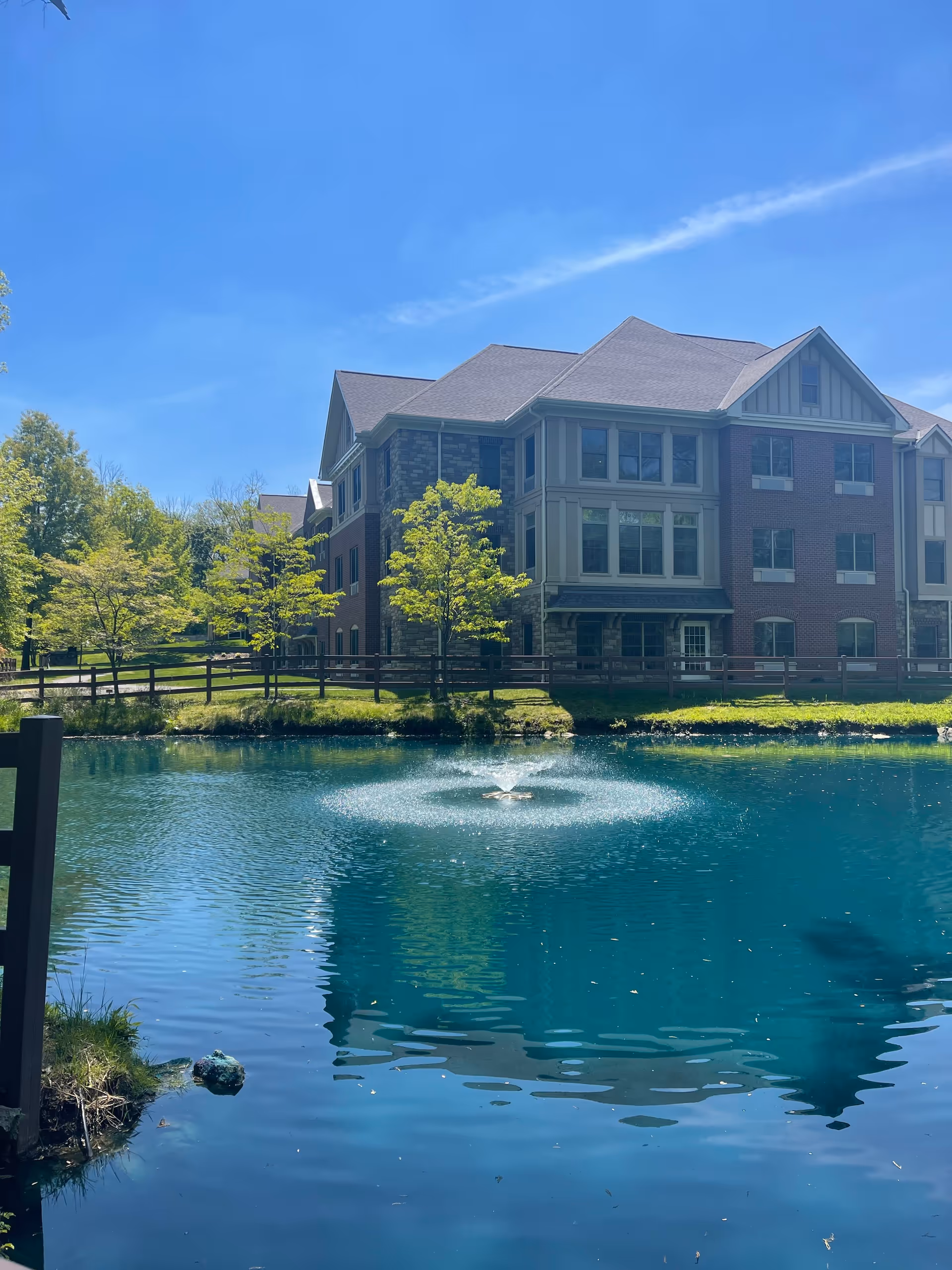 A multi-story brick and stone building with large windows is situated behind a small pond with a water fountain in the center. The pond is surrounded by green grass, trees, and a wooden fence under a clear blue sky.