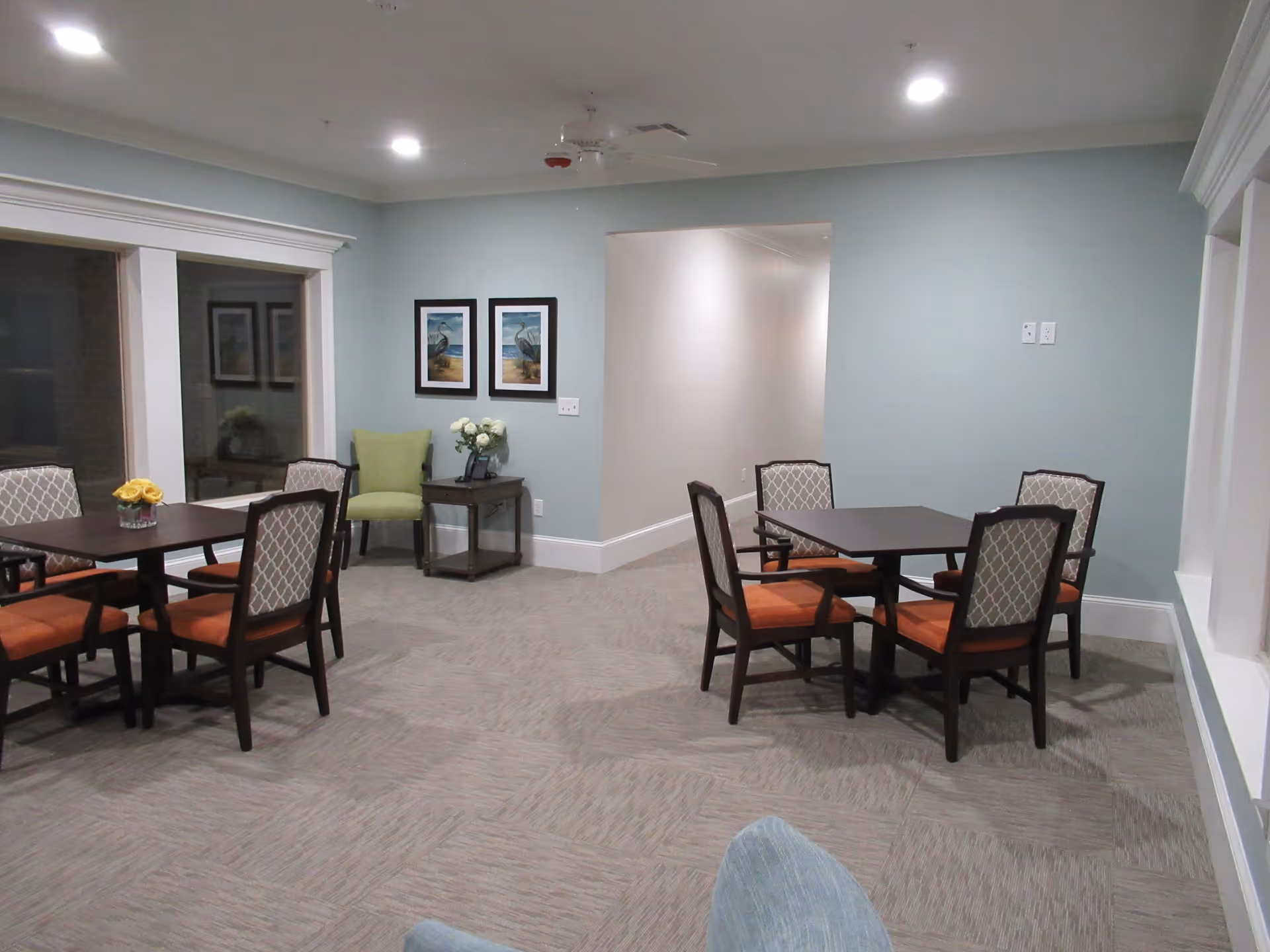 A well-lit interior room with two square tables, each surrounded by four chairs with patterned backs and orange cushions. There is a green armchair next to a small side table with a vase of white flowers and a phone. Two framed pictures of birds hang on the light blue wall. The floor is carpeted with a subtle pattern, and there is a large window on the left side.
