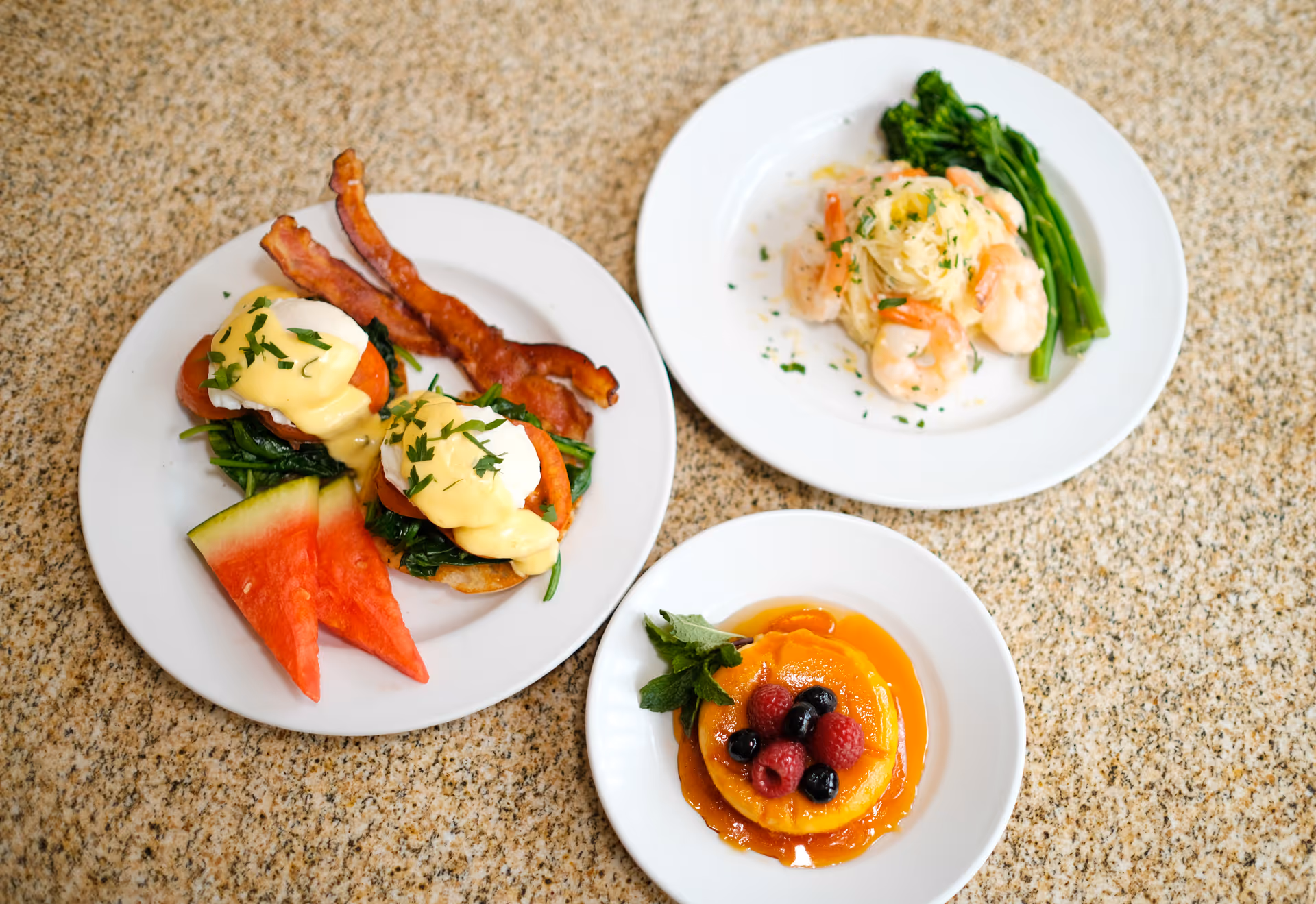Three white plates with eggs Benedict and bacon, a shrimp pasta dish, and a caramelized fruit dessert topped with berries on a speckled countertop.