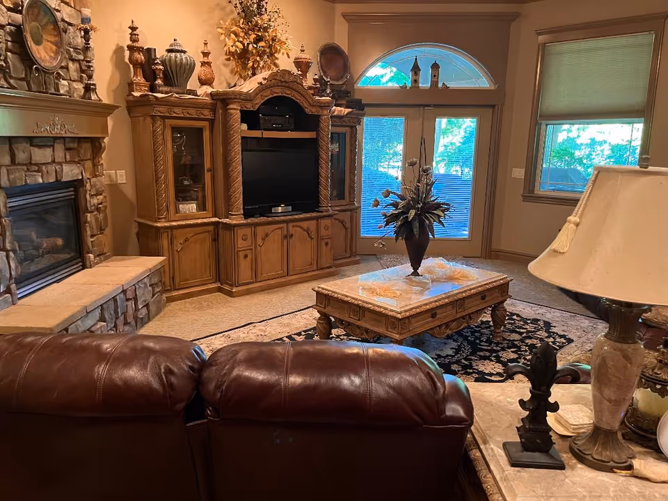 A cozy living room with a stone fireplace on the left, a large wooden entertainment center with a TV in the middle, and a glass door with an arched window above it letting in natural light. In the foreground, there is a brown leather sofa and a wooden coffee table with a floral arrangement on top. A table lamp and decorative items are on a side table to the right.