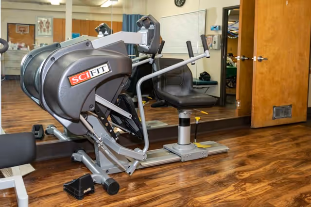 A fitness room with a SciFit exercise machine positioned on a wooden floor. The room has a large mirror on the wall reflecting the equipment and part of the room. There is a wooden door to the right and some medical or anatomical posters visible in the background.