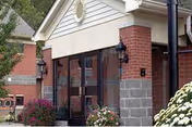 Exterior view of a healthcare facility entrance with brick and stone walls, glass doors, black lantern-style light fixtures, and surrounding landscaping including bushes and flowers.