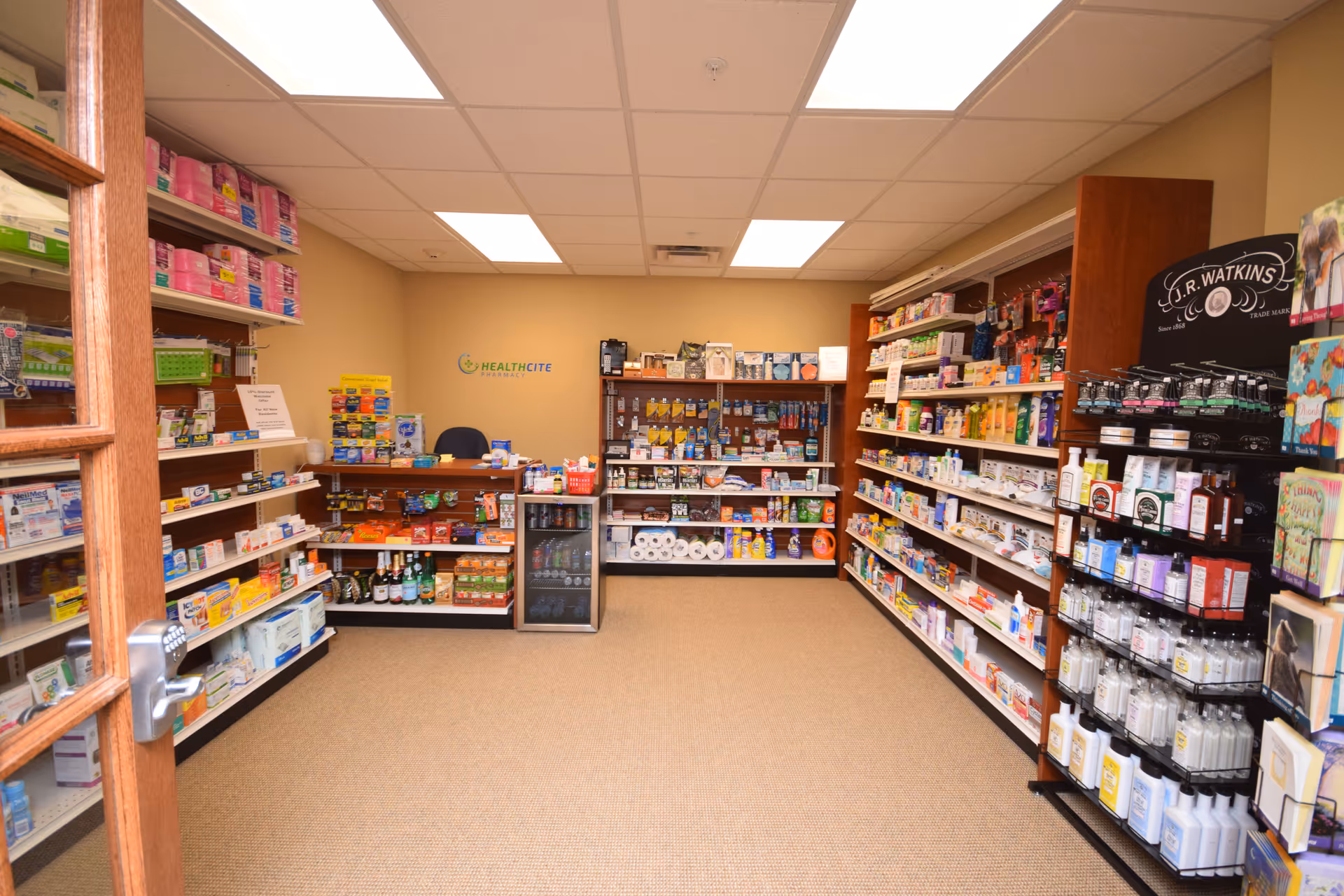 Interior view of a small retail pharmacy or convenience store area within a facility, showing shelves stocked with various health and personal care products, snacks, beverages, and toiletries. There is a counter with a chair behind it and a small refrigerator with drinks. The walls are beige, and the ceiling has fluorescent lighting.