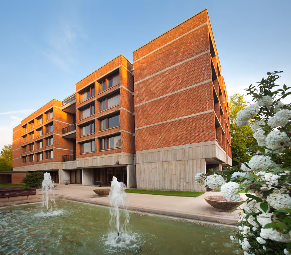 Brick multi-story senior living building with fountains and flowering shrubs in the foreground.
