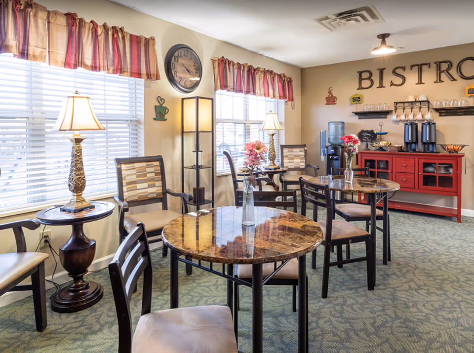 A cozy dining area with round marble-top tables and cushioned chairs. The room has two large windows with striped valances, two table lamps, and a floor lamp providing warm lighting. On the back wall, the word 'BISTRO' is displayed above a red sideboard holding coffee dispensers, cups, and a water cooler. Vases with flowers are placed on the tables.