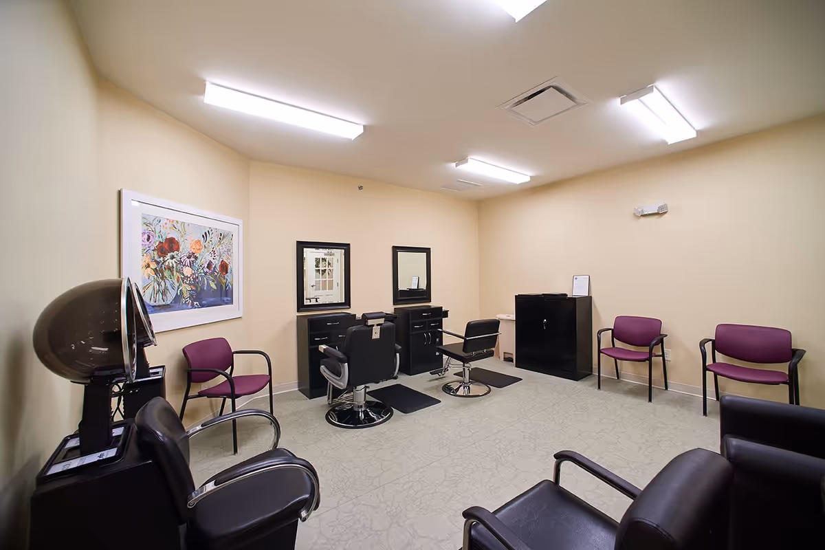 Interior view of a hair salon room in a senior living facility with two black salon chairs in front of mirrors and cabinets, several purple waiting chairs along the walls, a hair dryer station, and a colorful floral painting on the wall.