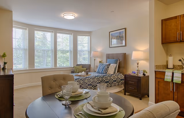 Sunlit studio apartment room with a neatly made bed, bedside tables and lamps, a round dining table set for two, and a small kitchenette.