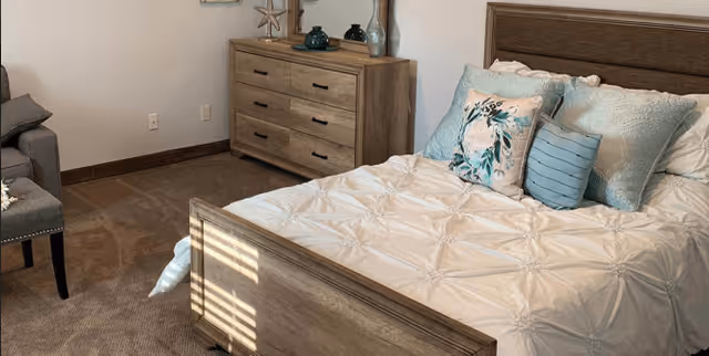 A cozy bedroom featuring a wooden bed frame with a white comforter and decorative pillows in shades of blue and floral patterns. There is a matching wooden dresser with decorative items on top, and a gray upholstered chair with nailhead trim is partially visible. The room has beige carpet and neutral-colored walls.