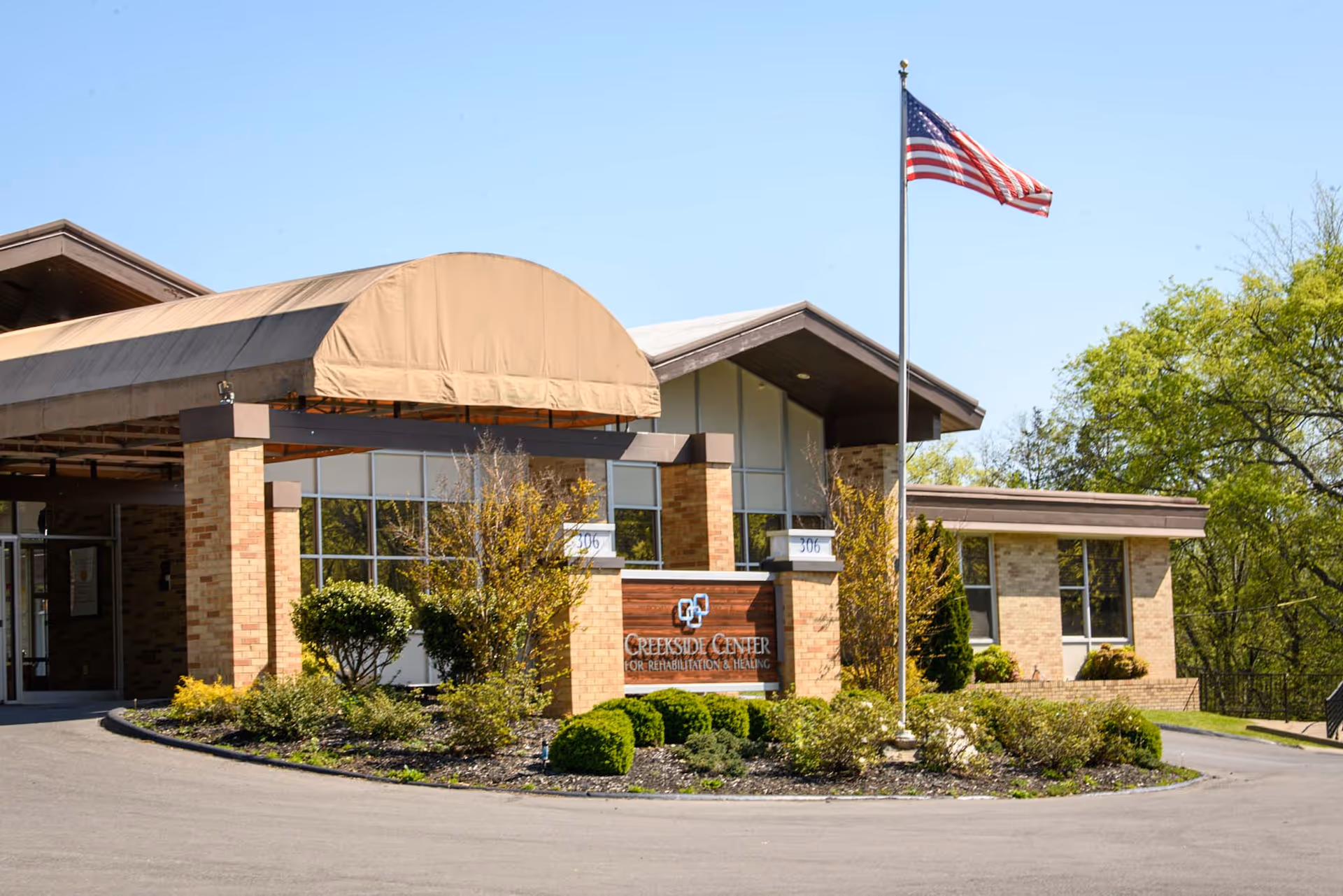 Exterior view of Creekside Center for Rehabilitation & Healing building with a covered entrance, brick walls, large windows, landscaped bushes, and an American flag on a flagpole in front.