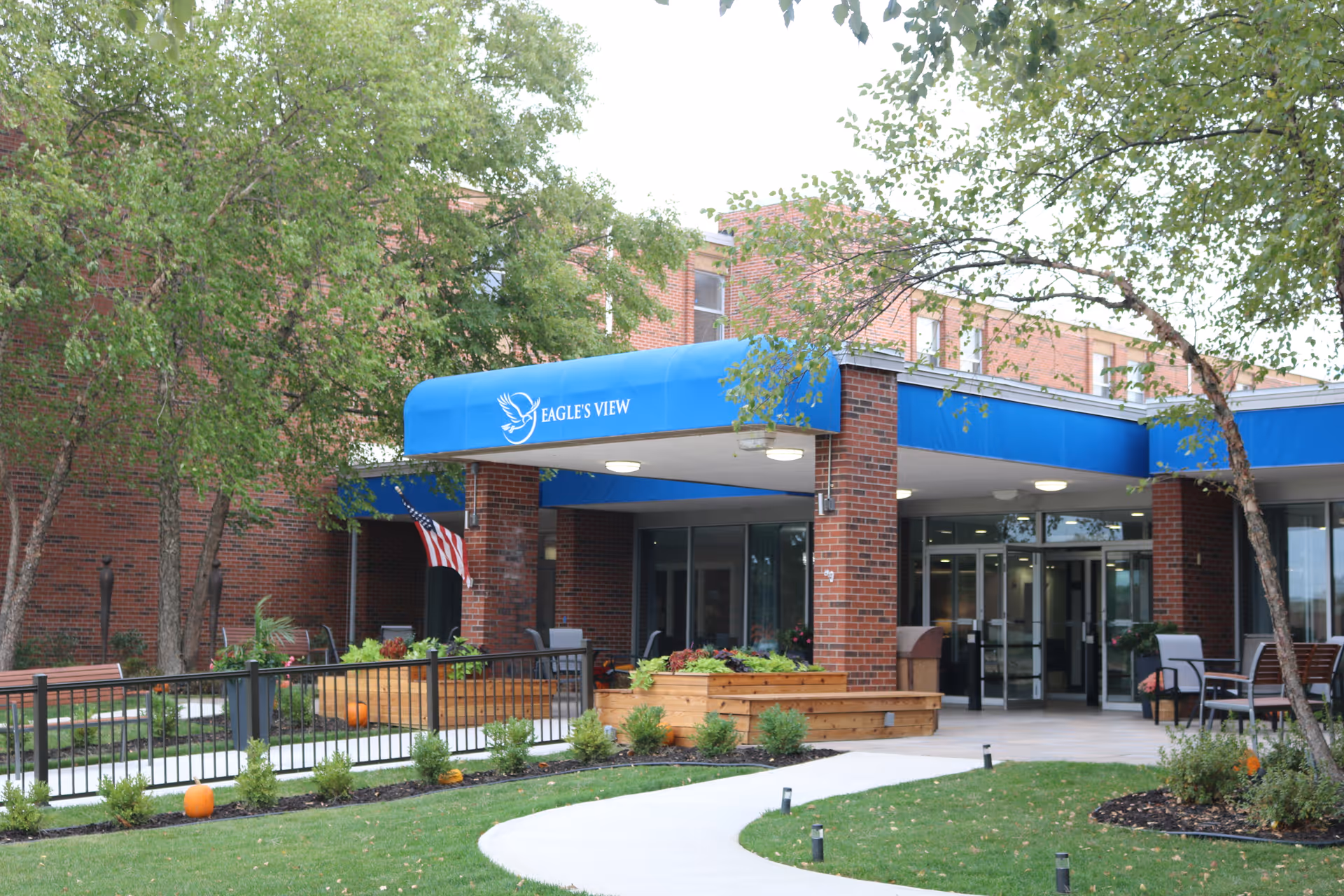 Entrance of Eagle's View Supportive Living and Memory Care facility with a blue awning displaying the facility's name and logo. The building is made of red brick and surrounded by trees, a well-maintained lawn, a curved concrete pathway, and outdoor seating areas with chairs and tables. There are also planters with greenery and pumpkins placed near the walkway.