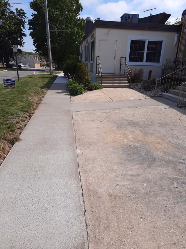 Sidewalk leading to the entrance of a single-story building with stairs and handrails, surrounded by some greenery and a small garden area. A sign on the grass reads 'Healthcare Heroes Thank You!'.