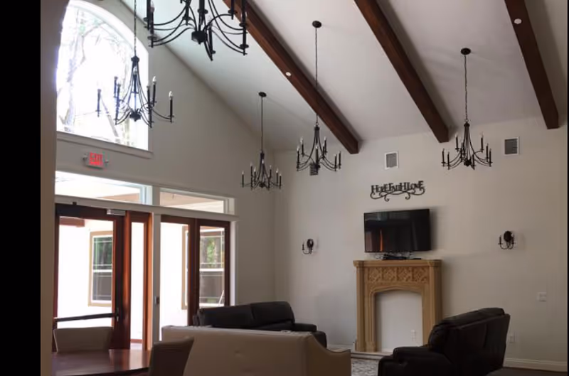A spacious living room with high vaulted ceilings featuring exposed wooden beams and multiple black chandeliers. The room has large windows and glass doors allowing natural light to enter. There are several sofas arranged around a decorative fireplace with a mounted flat-screen TV above it. The walls are painted white, and there is a decorative wall sign above the TV.