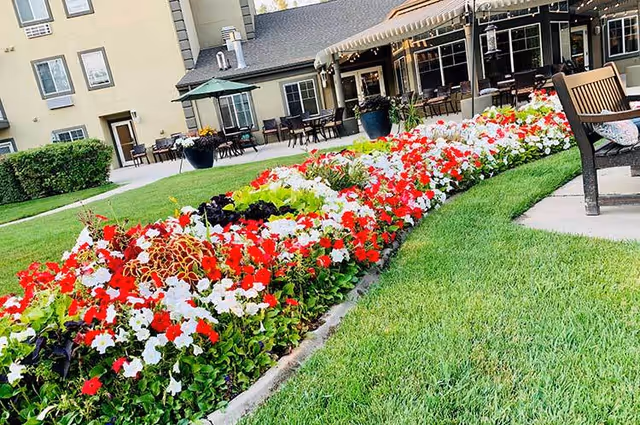 A vibrant flower bed with red, white, and green plants in front of a senior living facility building. There are outdoor tables with umbrellas and chairs on a patio area, along with a wooden bench on a grassy lawn.