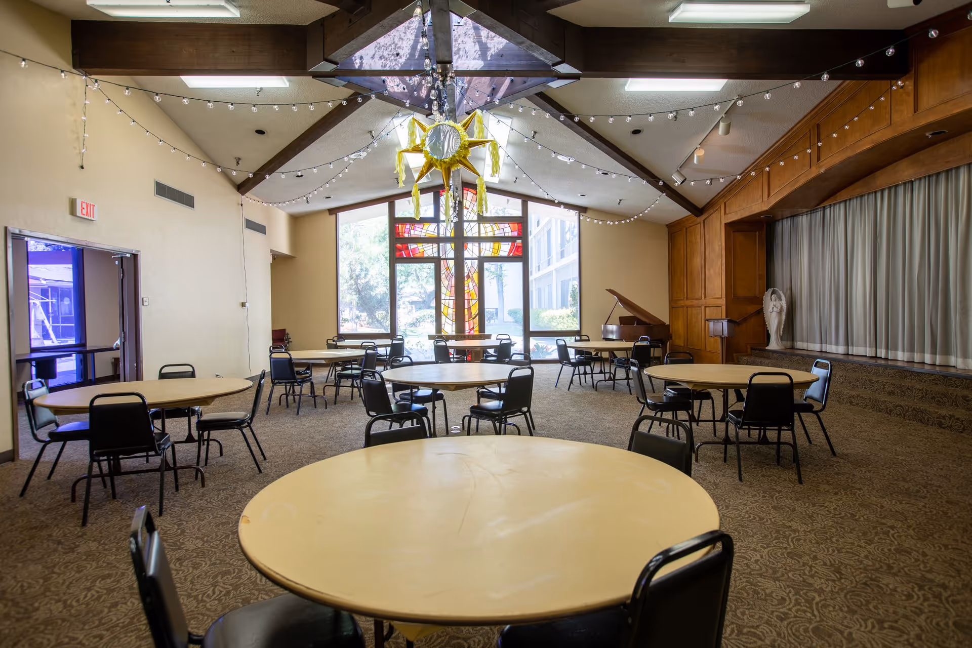 Interior of a community room with round tables and black chairs arranged on a patterned carpet. The room features a high ceiling with exposed wooden beams decorated with string lights. At the far end, there is a large window with a stained glass cross design, a grand piano, and a white angel statue near a curtained area.