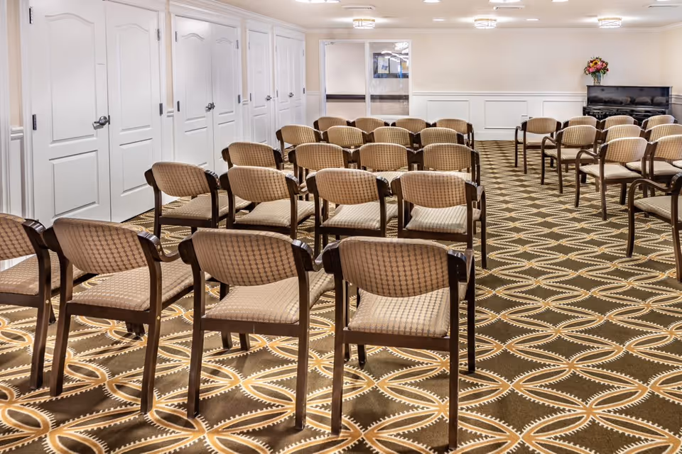 A room set up with multiple rows of beige cushioned chairs with wooden frames arranged facing a black piano with a vase of flowers on top. The room has a patterned carpet with circular designs, white paneled walls, and several white doors along one side.