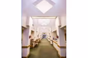 Wide, well-lit nursing home corridor with skylights and seating and tables along both sides.