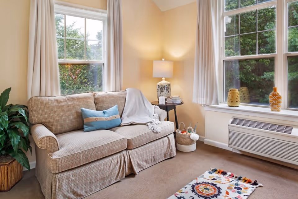 A cozy living room with a beige plaid sofa adorned with a blue decorative pillow and a light gray throw blanket. Next to the sofa is a small round wooden side table with a lamp, a mug, and some books. A woven basket with yarn and knitting needles is on the floor beside the table. Two large windows with white curtains let in natural light and show green trees outside. A colorful patterned rug is on the carpeted floor, and a green potted plant is placed to the left of the sofa.