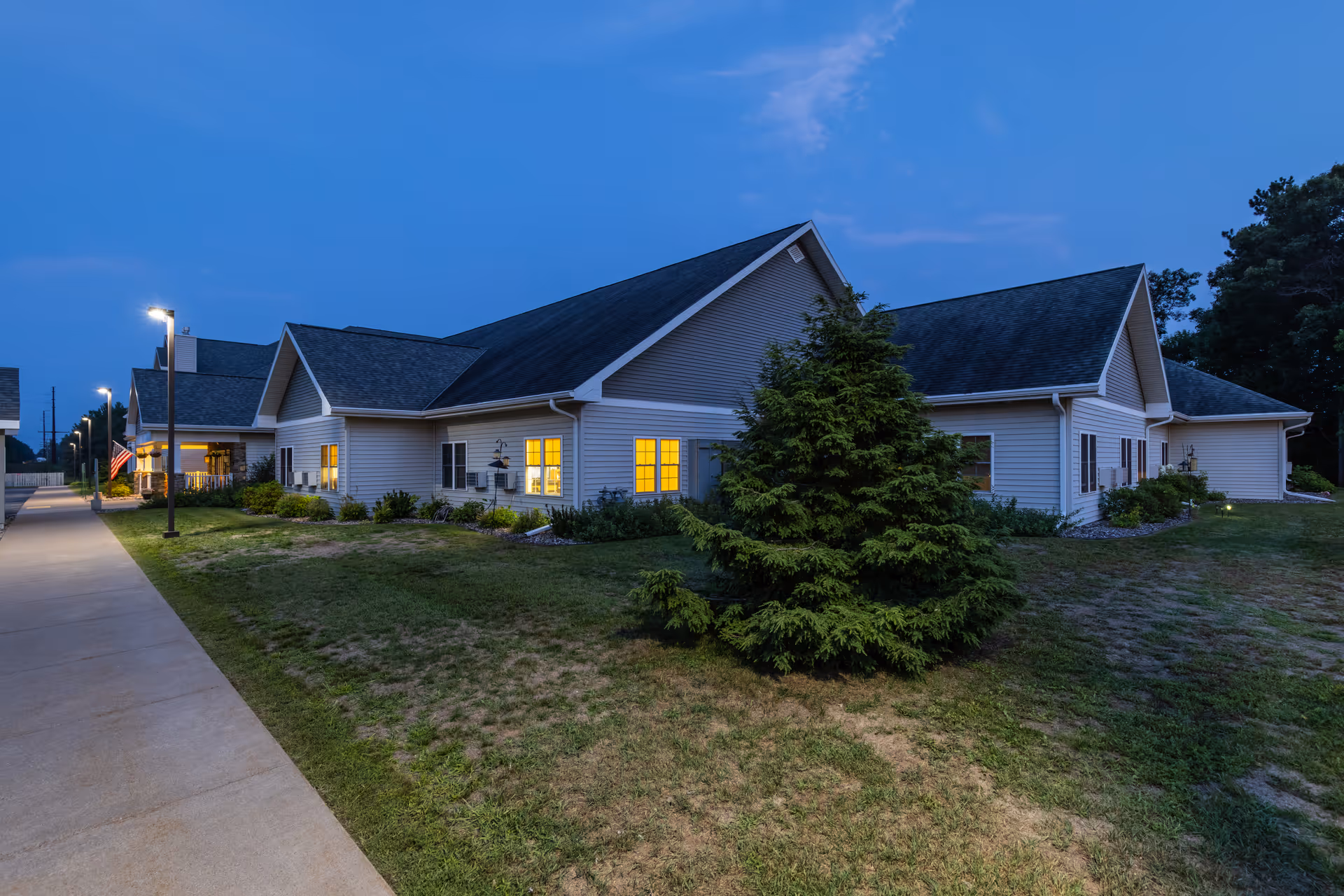 Exterior view of a single-story senior living building at dusk with lit windows, a sidewalk, and landscaped yard.