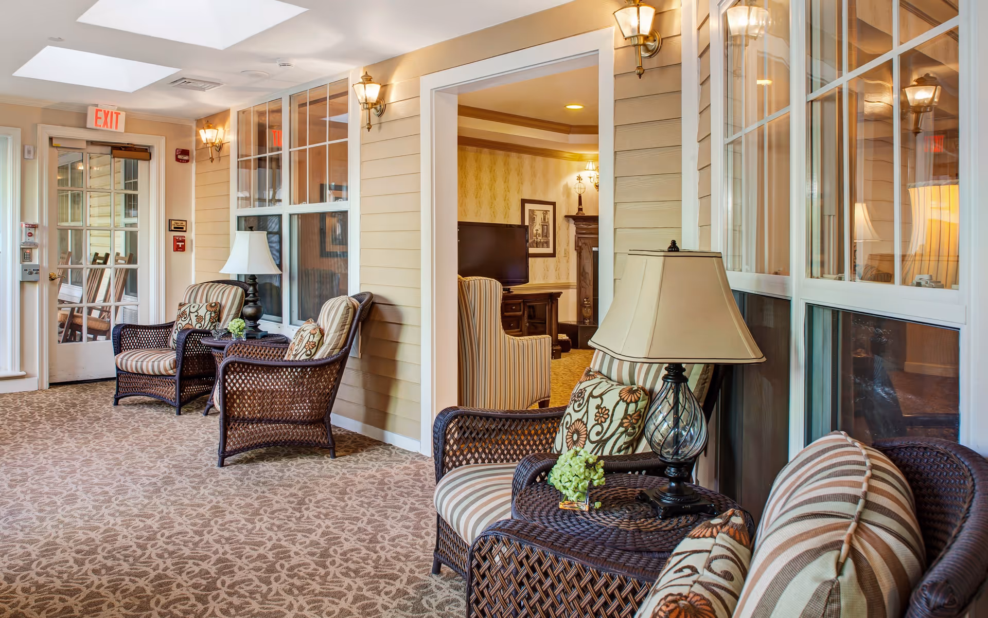 A cozy indoor seating area with wicker chairs featuring striped and floral cushions, small tables with lamps and a small flower arrangement. The area has beige walls, patterned carpet, and large windows. In the background, there is a view into a living room with a TV, armchairs, and framed artwork on the wall.