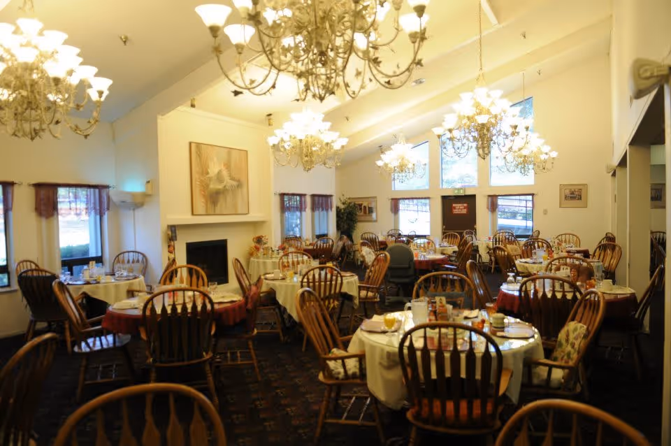 A spacious dining room with multiple round tables covered with white and red tablecloths, set with plates, glasses, and utensils. Wooden chairs with cushions surround the tables. The room features large windows with purple curtains, several ornate chandeliers hanging from a high ceiling, and a fireplace with a decorative piece above it.