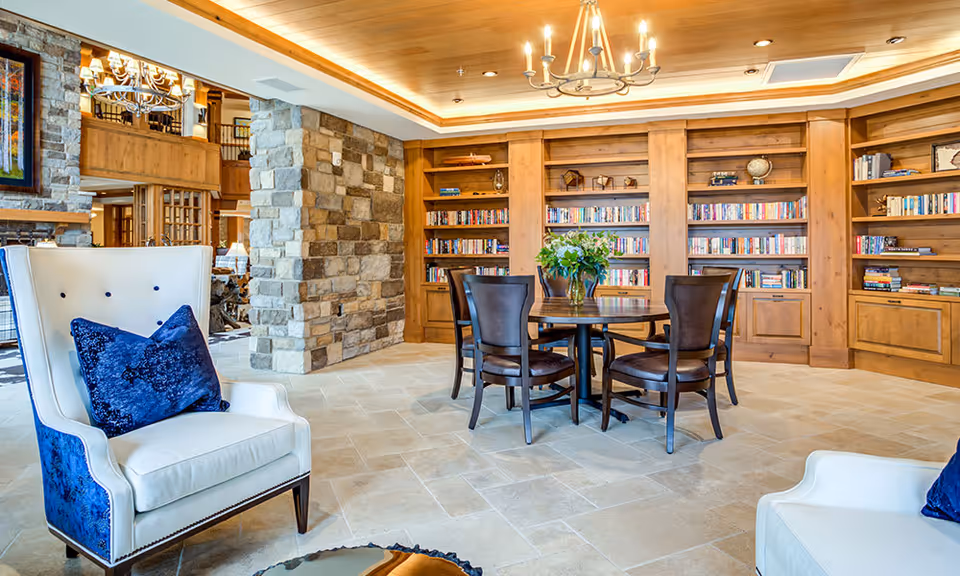 A cozy and elegant interior room featuring a round wooden table with four chairs, surrounded by built-in wooden bookshelves filled with books and decorative items. The room has a stone pillar, tiled floor, and a wooden ceiling with a chandelier. Two white armchairs with blue cushions are partially visible in the foreground.