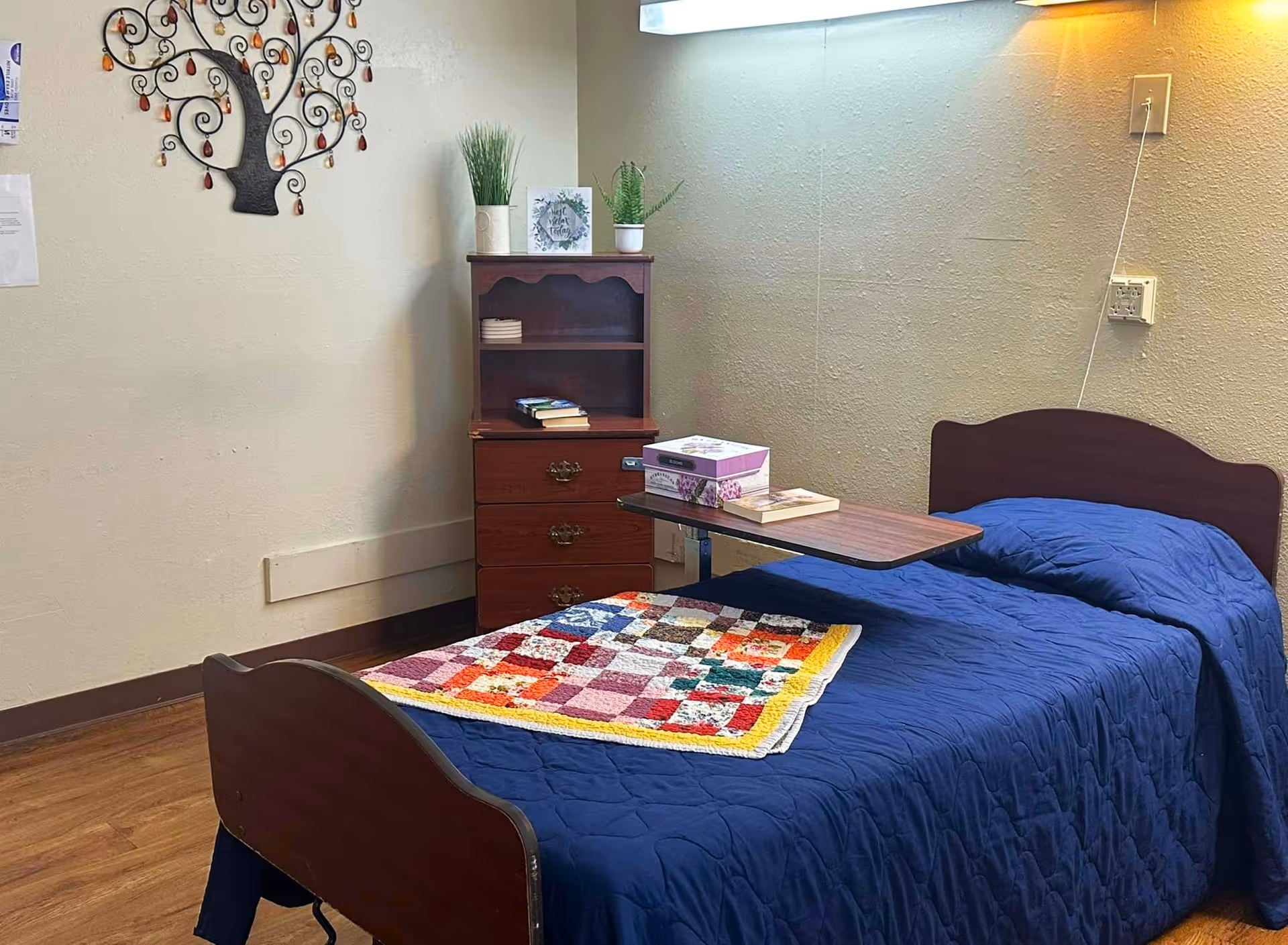 A small nursing center bedroom with a single bed covered in a dark blue quilt and a colorful patchwork blanket at the foot. Next to the bed is a wooden bedside table with three drawers and a small shelf on top holding books and two potted plants. A wall-mounted overbed table holds a box and a book. The walls are light-colored with a decorative metal wall art shaped like a tree and a fluorescent light fixture above the bed.