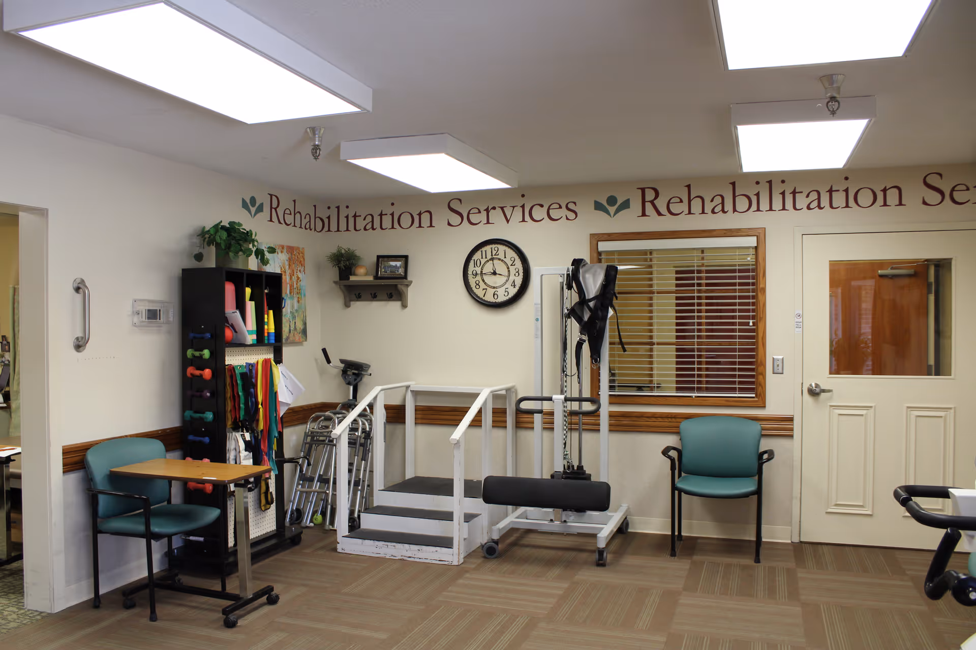 Interior view of a rehabilitation services room with exercise equipment including parallel bars and a harness system. There are two teal chairs, a small table, a black shelving unit with colorful exercise bands and small weights, a wall clock, and a window with blinds. The walls have the text 'Rehabilitation Services' partially visible.