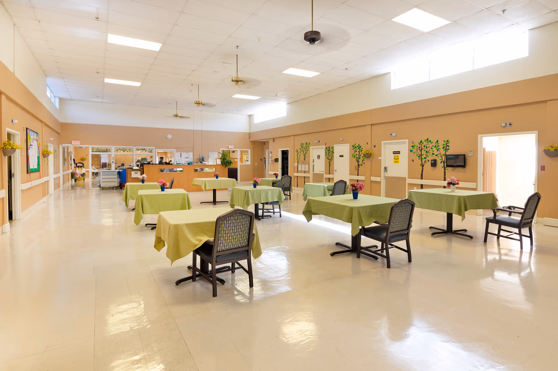A spacious dining area in a senior living facility with several tables covered in green tablecloths, each with a small flower pot centerpiece. There are chairs around the tables, beige walls with some wall decorations, and a reception desk in the background. The room has a high ceiling with ceiling fans and bright overhead lighting.