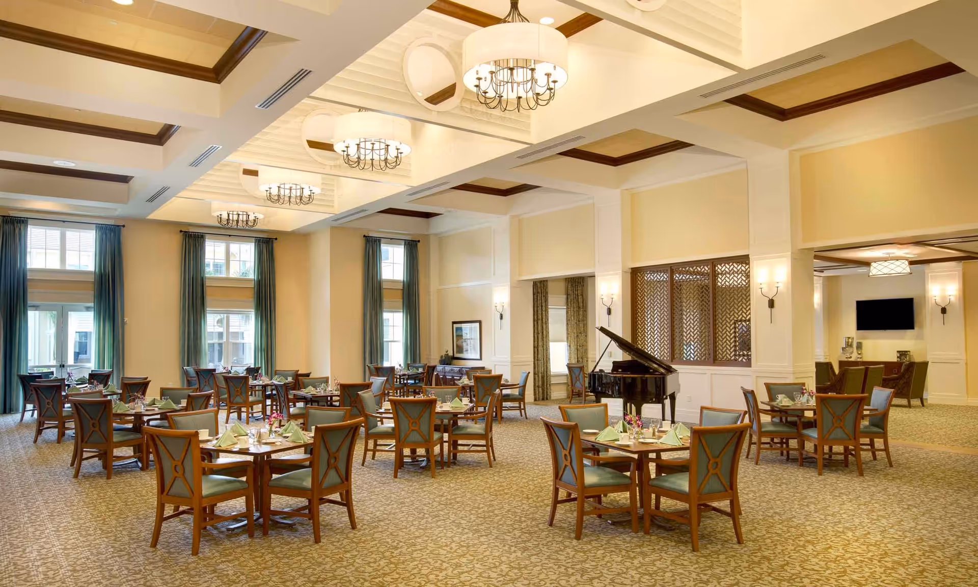 A spacious and elegant dining room with multiple round wooden tables and chairs arranged neatly. Each table is set with green napkins and small flower arrangements. The room features large windows with green curtains, a grand piano, and warm lighting from chandeliers and wall sconces. The ceiling has decorative beams and recessed lighting.