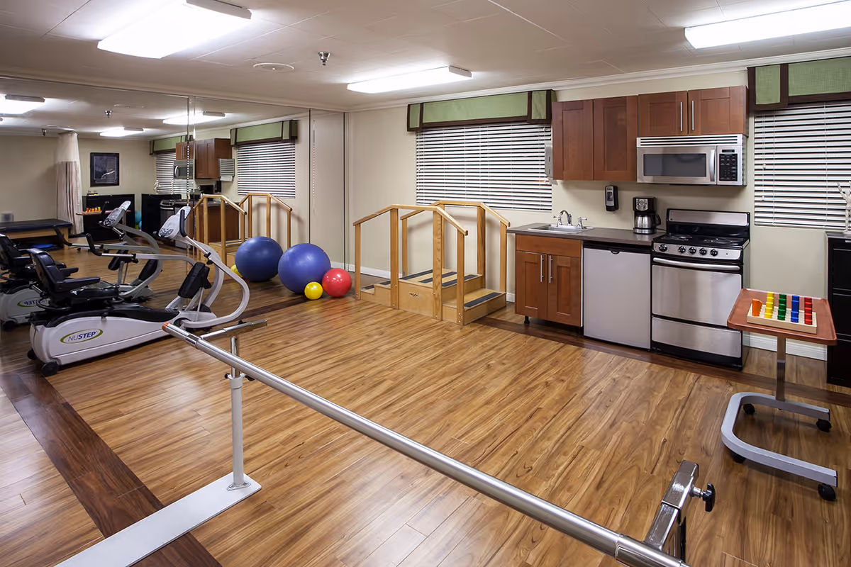 A rehabilitation room with exercise equipment including stationary bikes, therapy balls, parallel bars, and a small set of stairs for physical therapy. The room also features a kitchenette area with wooden cabinets, a sink, microwave, stove, and mini fridge. The floor is wood laminate and there are large windows with blinds.