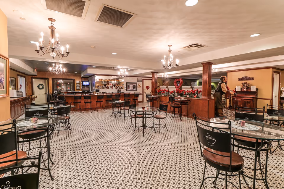 A spacious dining area with multiple round glass-top tables and black metal chairs with wooden seats. The floor has a black and white patterned tile. There are several chandeliers hanging from the ceiling providing warm lighting. In the background, there is a bar with high stools and a counter, decorated with heart-shaped ornaments and red flowers. A statue of a man playing an accordion is visible near the right side of the room.