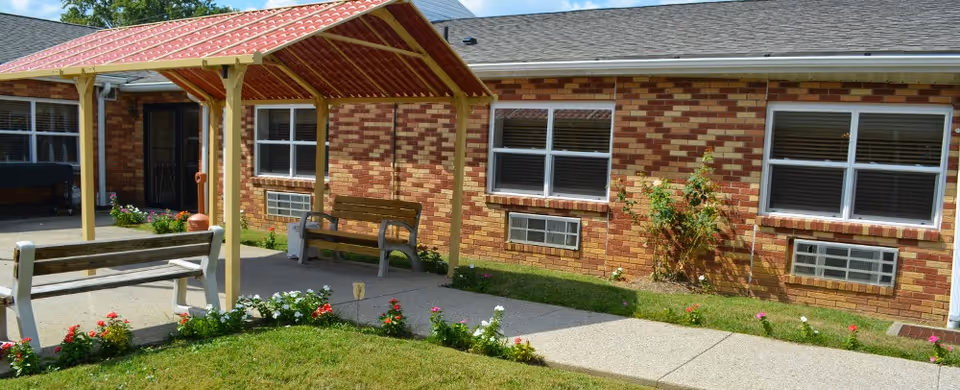 Outdoor seating area at Good Samaritan Home and Rehabilitative Center featuring a small covered structure with a red roof, two wooden benches, a concrete pathway, and a garden with flowers in front of a brick building with multiple windows and air conditioning units.